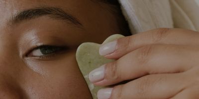 Part of a woman's face is visible. She is staring at the camera and holding a green gua sha to her left cheek, pulling it away. She has a white towel on her head.