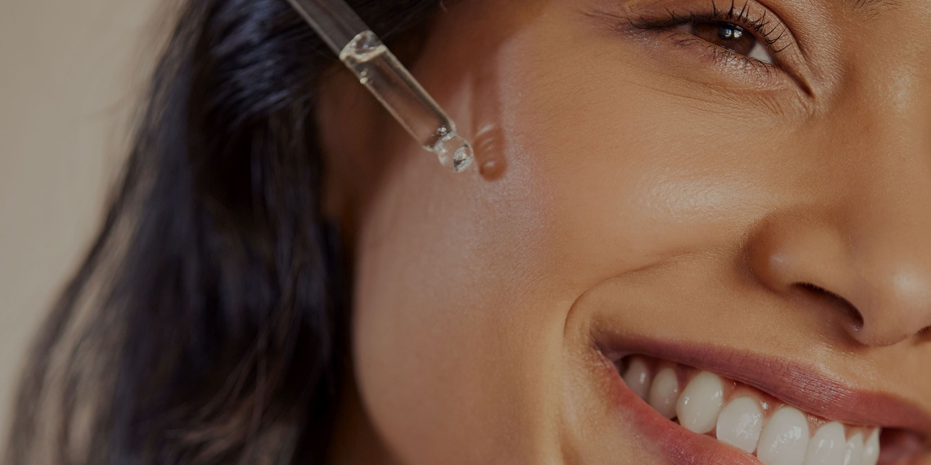 An image of a woman smiling with a serum dropper next to her cheek